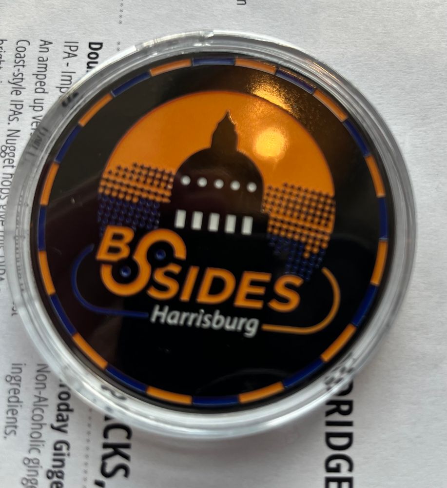Metal coin in a plastic case. Alternating blue and yellow dashes along the edge. Blue and yellow BSides Harrisburg logo in the center.