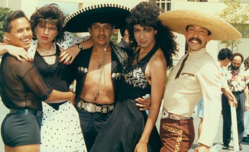 Pictured: Colored photograph of five people. From right to left, a person with a short haircut, black mesh t-shirt, shiny black belt, and black booty shorts with their right arm wrapped around another person with a bob haircut, red lipstick, a cropped black and white polka dotted jacket with a matching skirt, a black shirt, and red nail polish. They have their right arm wrapped around the leftmost person, and the left arm on the shoulder of a person with a short haircut wearing a black sombrero, an open jacket with silver embellishments, a large embellished belt, and black pants. This person is holding the person to their right and their left hand wrapped around the waist of a person with long, curly hair, a large black beaded necklace, a black halter dress with sequined cups. Standing on the left of them with a straw sombrero, thick moustache, a tan shirt with a brown tie, an embellished belt, and embroidered brown pants. Each member of the group looks directly at the camera, smiling.