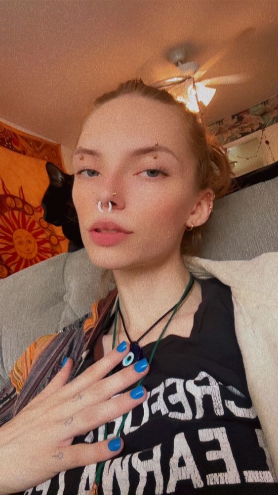 Violet sits on the couch. Their hand placed in their chest and nails pained blue poses for the camera 