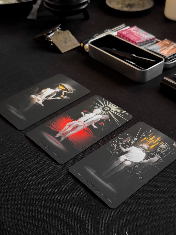 Three modern tarot or oracle cards laid out on a black cloth surface, each featuring artistic black and white photography with colorful accent elements - one with yellow/gold, one with red, and one with dramatic lighting effects. A metal tin case containing additional cards and supplies sits in the background, along with what appears to be a vintage flask.
