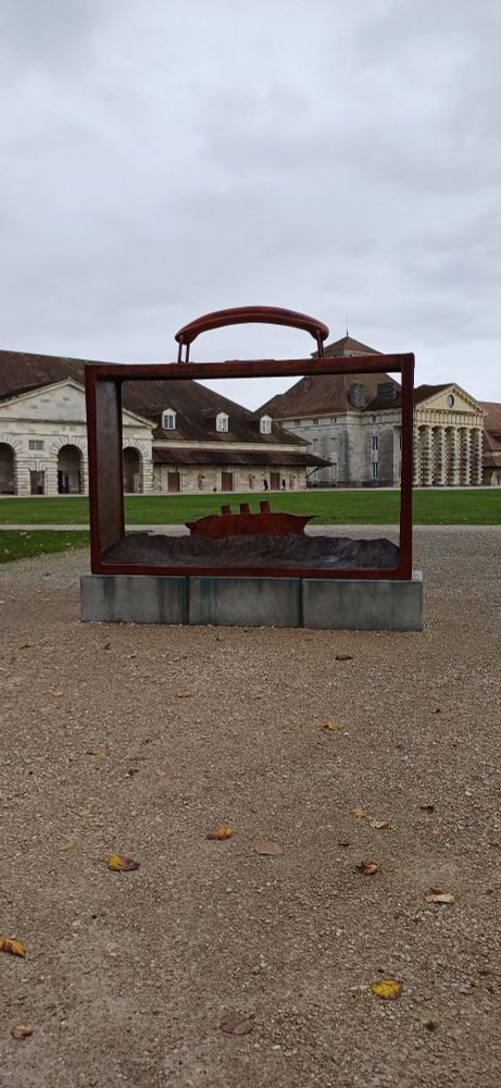 Statue de Folon : une valise évidée avec un bateau à l'intérieur. A l'arrière les bâtiments de la Saline Royale d'Arc-et-Senans