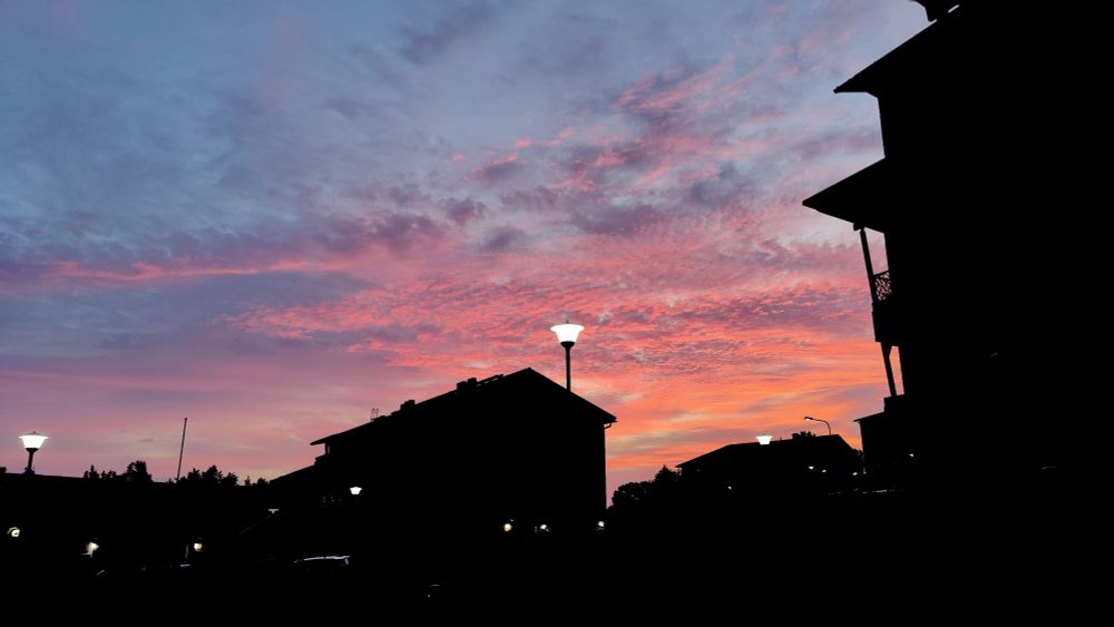 Photo of the sky at 4 AM. The clouds have taken on hues of orange, red, pink and purple from the rising sun (not pictured.) The buildings in the foreground have been darkened to completely black silhouettes.
