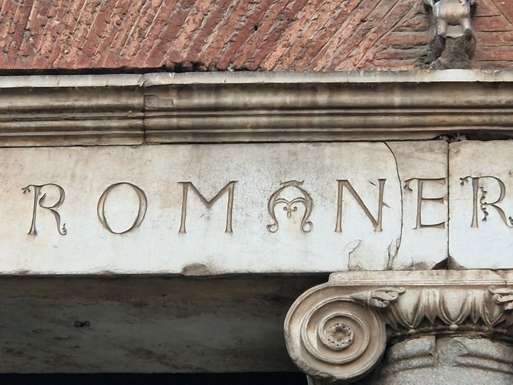 A frieze with the word "Romane" inscribed on it. The A is shaped like a woman's face. 