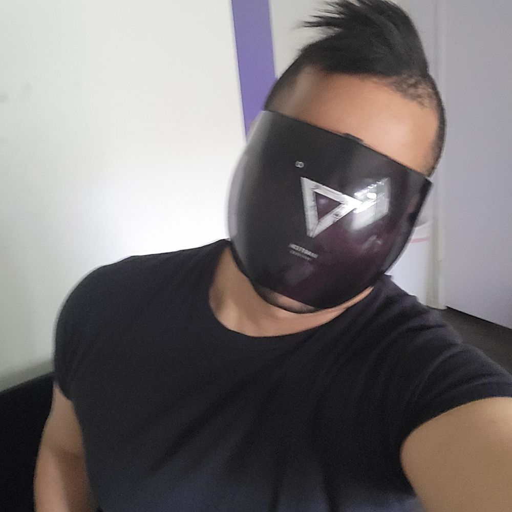 Cade in a black face mask with an upside down triangle in it. He's wearing a black shirt and his face is concealed. 
