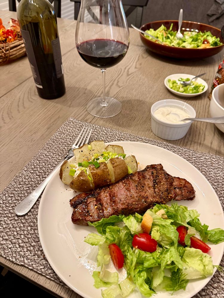 Perfectly grilled steak and cut baked potato loaded with butter, sour cream, and chives. Light green romaine salad with cherry tomatoes and croutons. All on a white dinner plate, brown and white placemat on the table. Serving dishes visible behind along with a dark bottle of Cab Sav and generous amount in a rather large, thin, Pinot glass. 