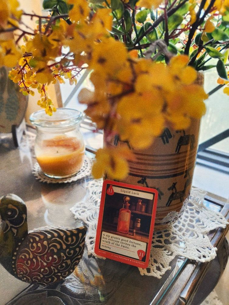 A photograph of a side table with a yellow wide-base candle in the background, a large vase with yellow flowers on a white doilie, an orange card titled 'Liquid Luck' rests against it as the focal point of the image. An ornamental duck sits off to the edge of the frame.