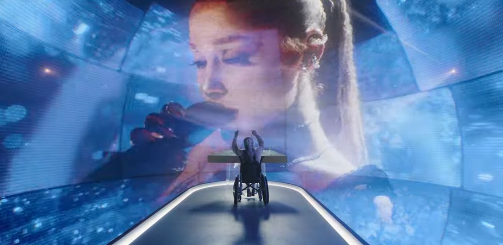 Older version of Ariana Grande in a wheelchair positioned on a futuristic, illuminated pathway with their arms raised toward a large digital screen. The screen displays a close-up of her younger self holding a microphone, with a soft blue-toned background creating an immersive concert-like atmosphere
