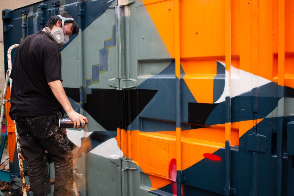 A man in black sprays an orange spray can to his side in preparation to continue painting the metal storage containers in front of him.