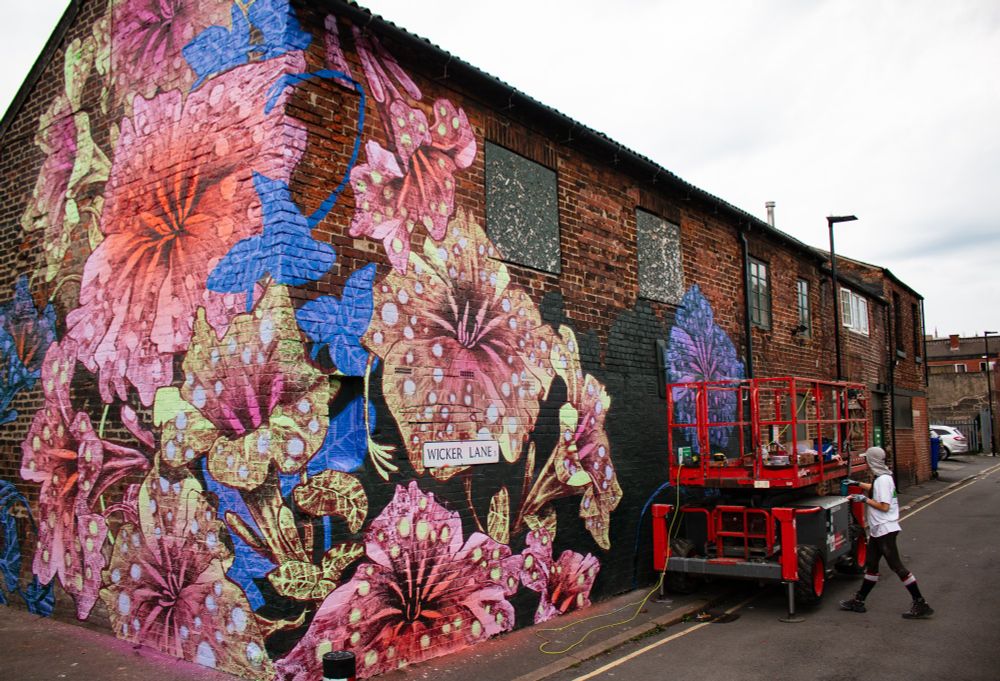 A pink and green painting of giant flowers on the side of a building. A scissor life on the right with the painter standing next to it.