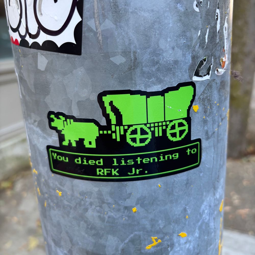 A lime green sticker on a metal pole. It’s pixel art from the original Oregon Trail game that reads “You died listening to RFK Jr.”