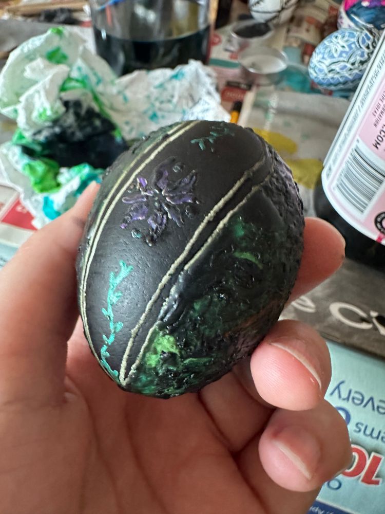 Larysa holds a wax covered pysanka delicately between their fingers. Beneath the murky dark wax you can see white lines down the curvature of the egg, as well as teal vines, a purple flower, and a large swath of greens.
