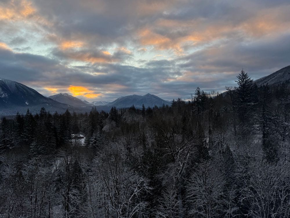 Sunrise photo from a high rise, showing snowy trees, mountains, and some orange and yellow light in the clouds