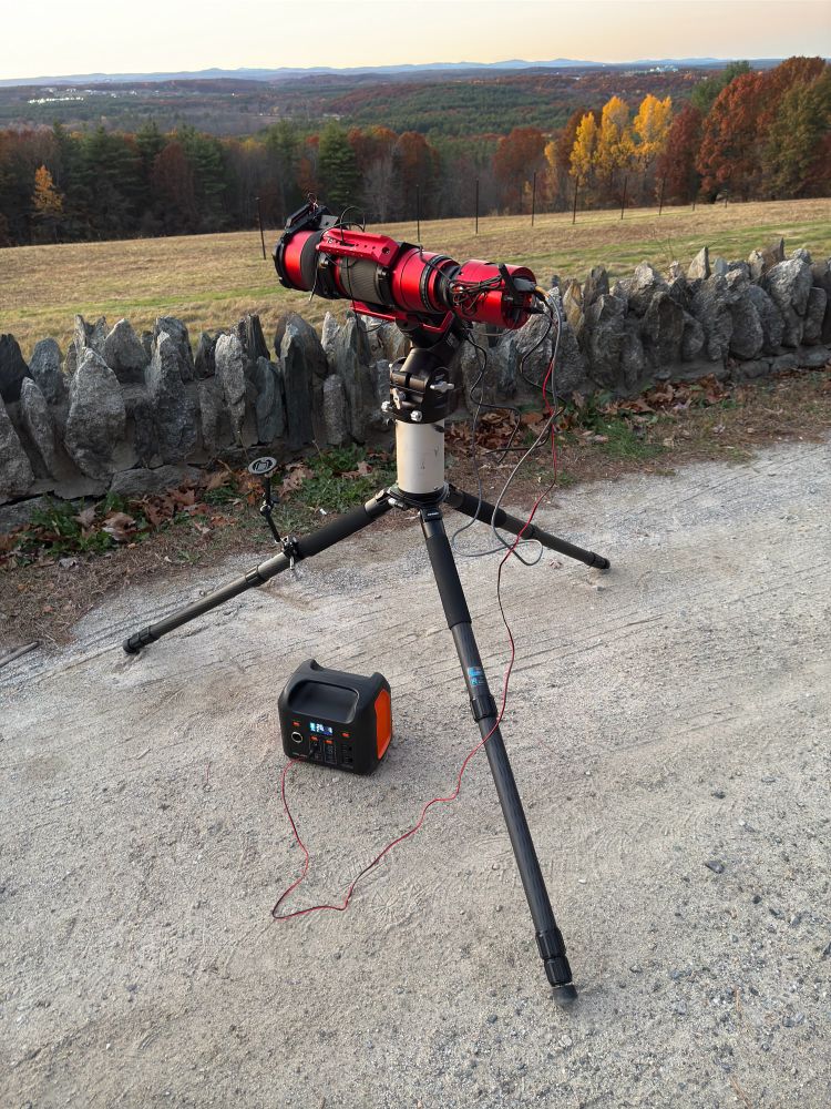 Telescope on a tripod sitting by a wall looking over a field and trees. There is a battery underneath connected to the telescope. 