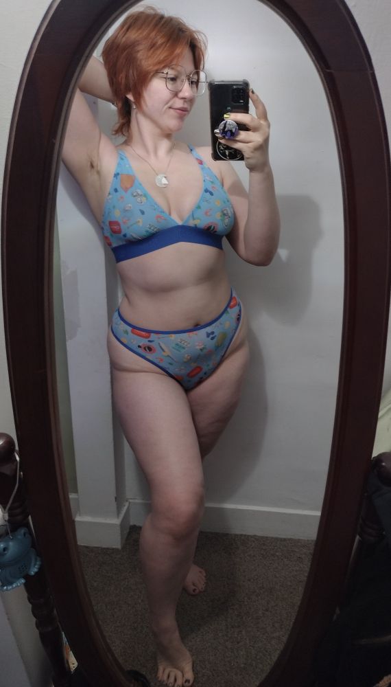 mirror selfie red head woman wearing square frame glasses and blue mesh triangle bralette with matching high waisted thong underwear and silver crescent moon and triangle pendants on silver necklace chain