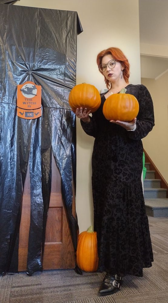 woman red shoulder length red hair oversized glasses holding two fake pumpkins wearing long black velvet dress next to a sign reading "the witch is in"
