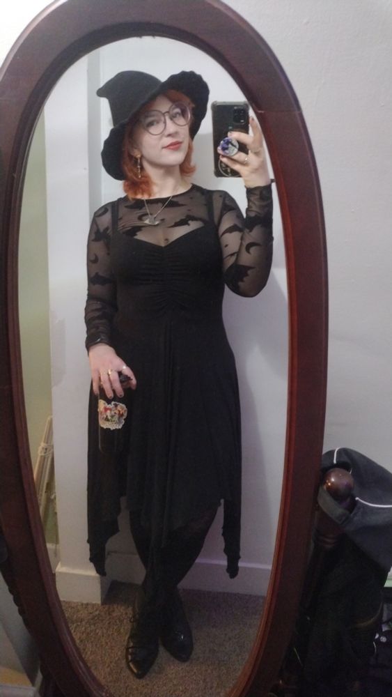 mirror selfie woman red shoulder length hair witch hat oversized glasses mesh bat print long sleeve black handkerchief hem dress black boots holding coffee thermos