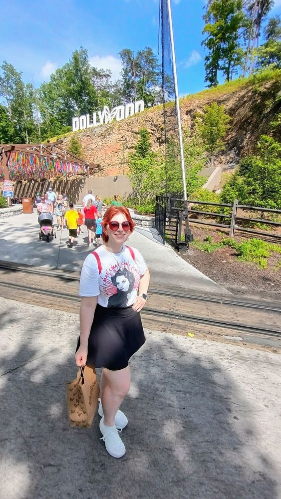 woman with red hair poses in front of Dollywood sign wearing sunglasses a white CMAT concert t-shirt black skort and white trainers holding a paper Dollywood branded bag