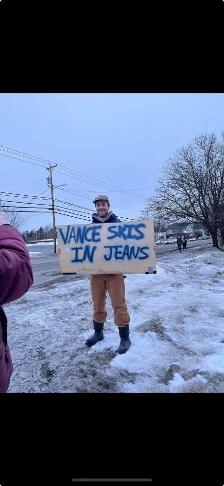 photo of a protestor standing in the snow holding a sign that says "Vance skis in jeans"