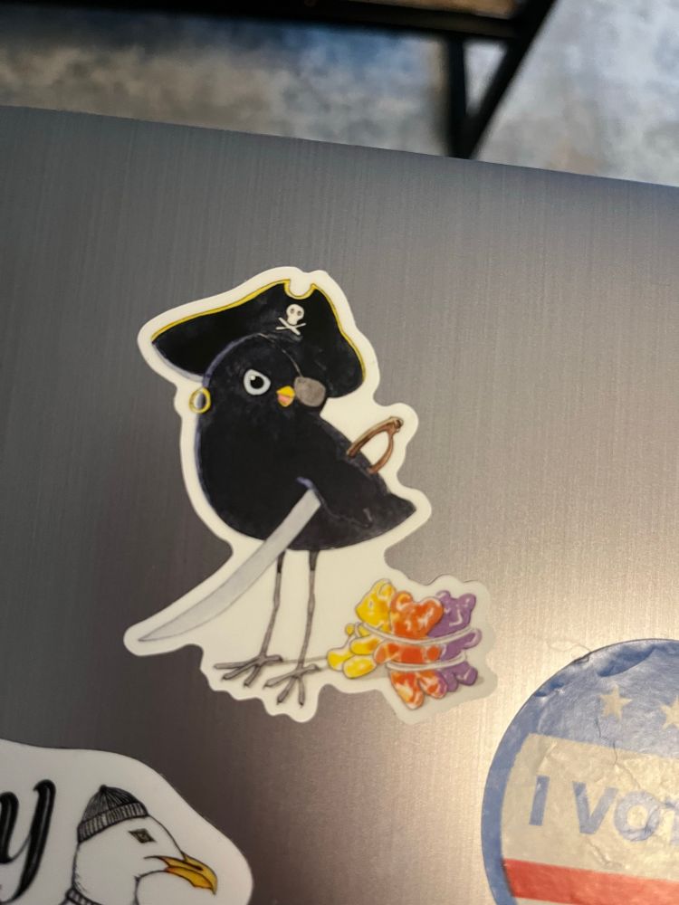 Sticker on a laptop that shows a small black bird wearing a pirate hat, an eyepatch and a gold hoop earring. A sword is tucked under its wing. Tied to its leg are three gummy bears bound in rope as if they’d been captured by the fiercest pirate in the skies.
