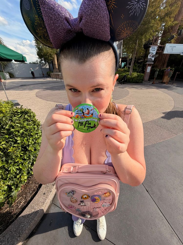 Danielle holding a pin that says “I’m Celebrating 1st Pin Trade” up to her face with a backpack displaying various pins (peep the hmj pin on my backpack)