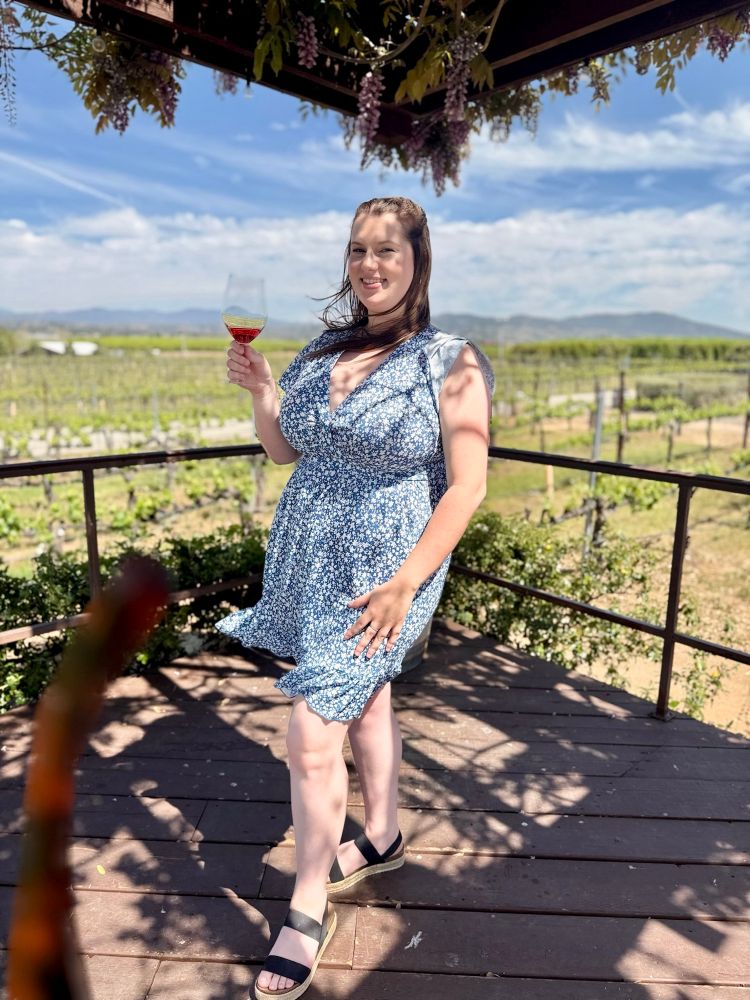 Danielle standing with a glass of wine with scenic vineyards and mountains in the background