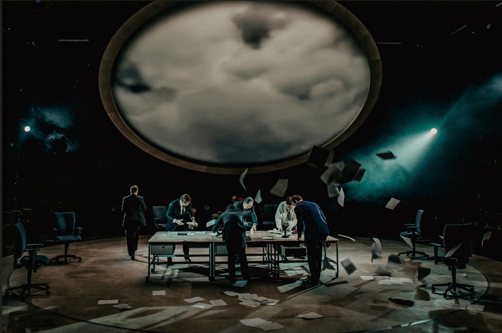 an image from the play "Agreement": a group of people at a table shuffle papers as papers seem to blow around them in a storm