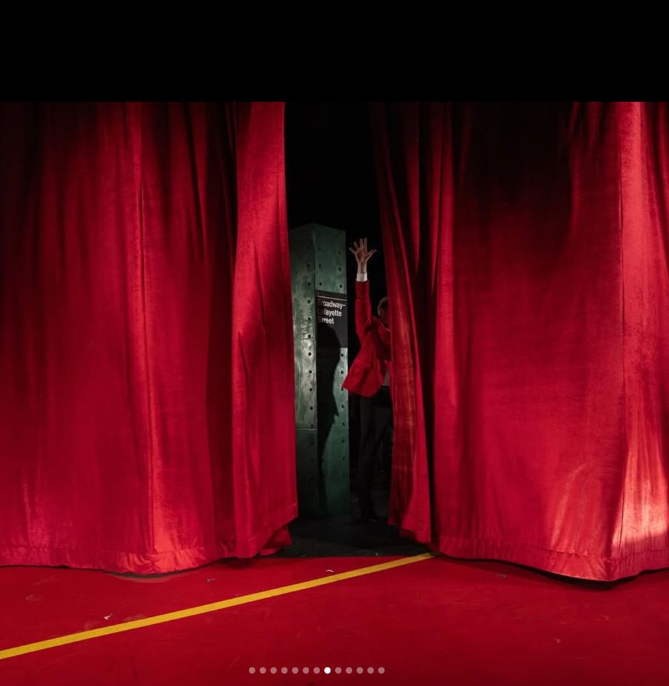 A red curtain closes on a red-painted stage. In the gap between the curtains, there's a green column from the subway, with its sign for "Broadway-Lafayette", and a man in a red jacket in waving to (or pushing?) someone, but the curtain prevents us from seeing which