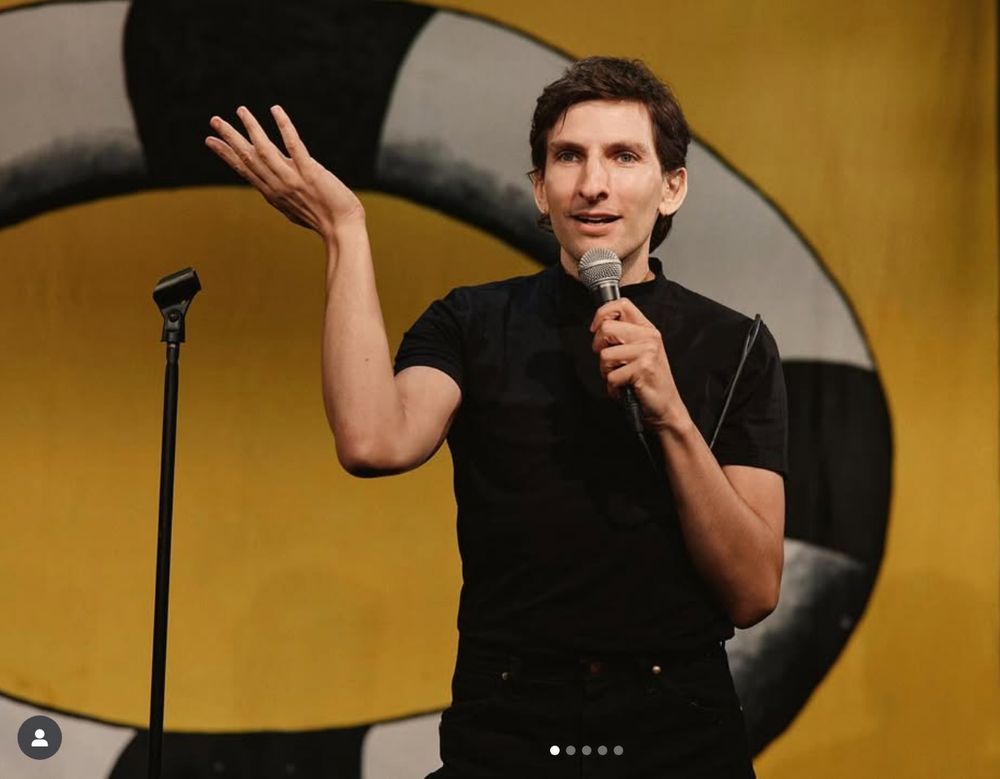 A tall, dark haired performer in a black tshirt gestures magnificently while speaking into a microphone. 