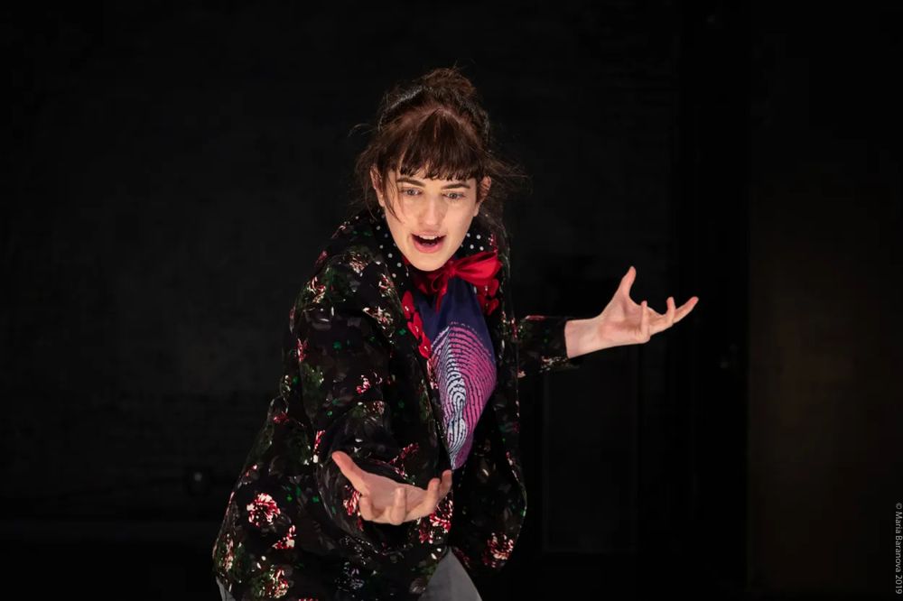 A woman dressed as a magician in a sparkly frock coat gestures magnificently