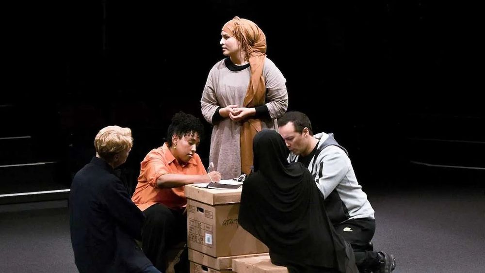 an image from the play "Grenfell": a group of four people sit around a pile of cardboard boxes filling out forms on the box-lid; a woman in a headscarf stands behind them