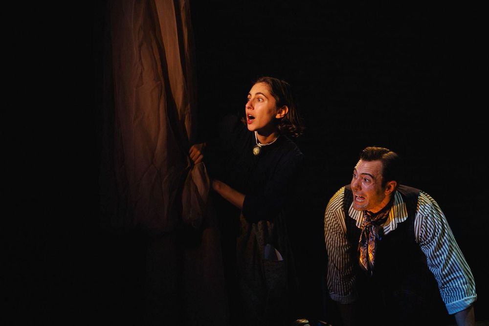 Madeline Weinstein and Neil D'Astolfo are clearly making Cockney noises while staring, shocked, from behind a curtain. They could be lit by candlelight.