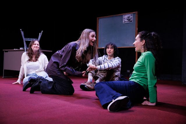 Four high school students sit on a red carpet with a chalkboard behind them; in the front, a girl in plaid shirt and engineer boots seems to be saying something urgently, and the other three stare at her, hypnotized