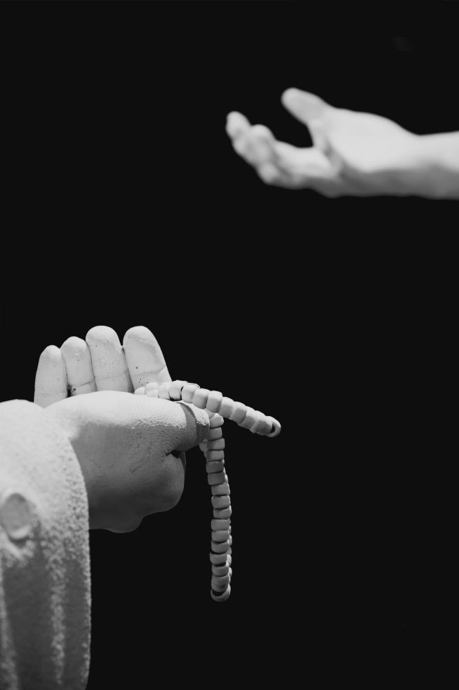 A high contrast black and white image of a statue hand offering a necklace of beads to another statue hand