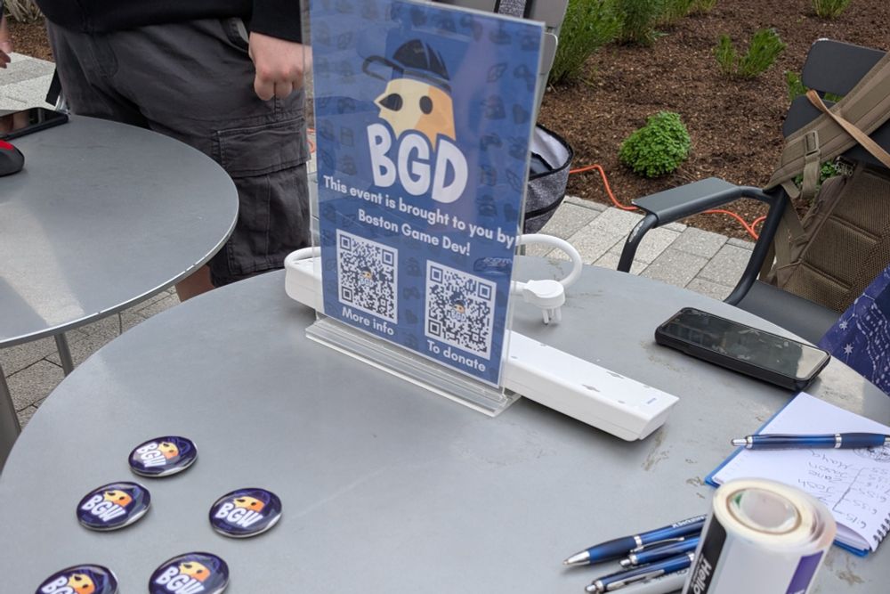 A table with several BGW pins, name tags, and a poster that says "this event is brought to you by Boston Game Dev!"