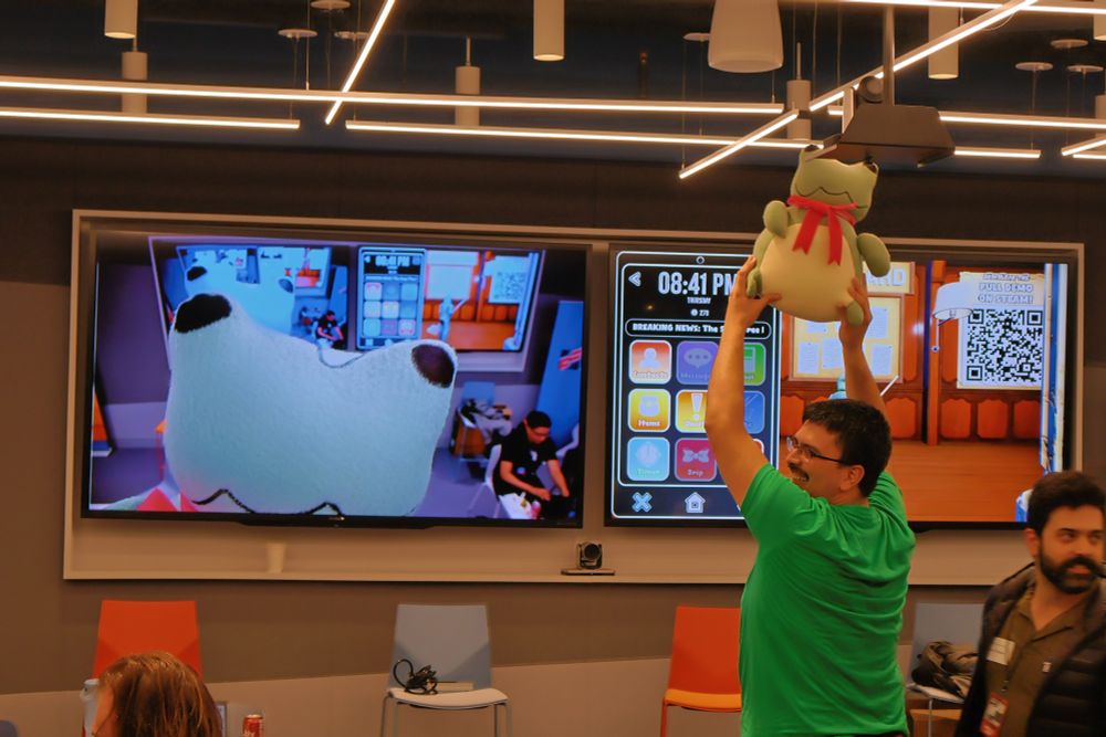 A photo from Made in MA. Manny Mallea is holding a frog plushie from the game Frogmageddon in front of a mounted camera. Its big frog face appears on the large screen behind Manny.
