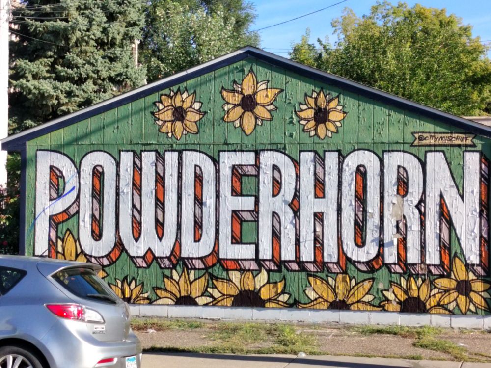 Photo of a mural that says "Powderhorn" on the side of a garage in morning sunlight. The word is written in big, white all-caps in a nice font with multicolored 3D edges, on a green background surrounded by paintings of sunflower blooms. The sunlight is coming from the side, highlighting the texture of the old vertical wood siding and where the paint of the mural is starting to peel. If you're familiar with the neighborhood you may recognize that this is on 31st Street just west of Bloomington Avenue.