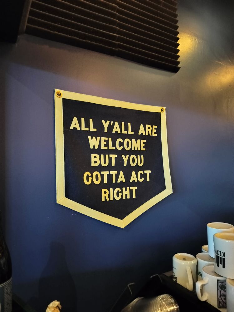 Canvas banner, like the ones moons posted to begin with, that says, "All y'all are welcome but you gotta act right." This one is hanging at the end of the bar at a coffee shop and brewery, you can see a stack of coffee mugs.
