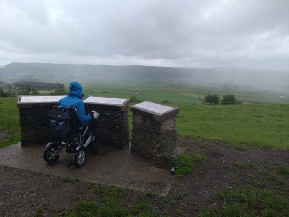 Rainy Cumbria hills. Sorrel is in their powerchair with their back to the camera, waterproof hood up, looking at the viewpoint maps of the hills. Probably to no avail, given the mist and general lack of visibility. 