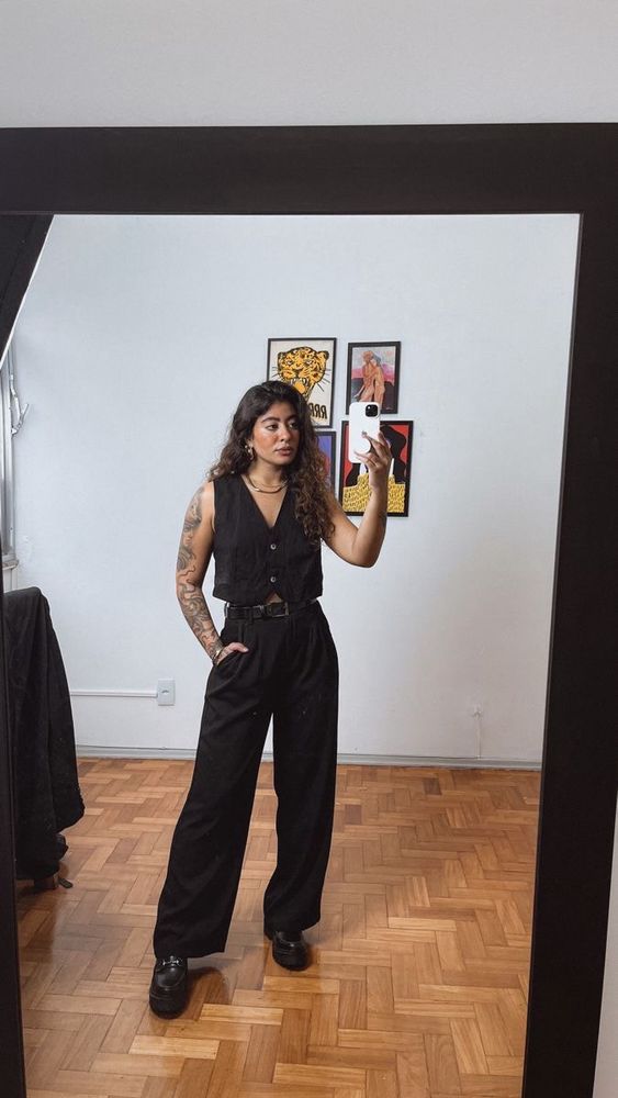 person with long, curly hair and tattoos taking selfie. They are wearing a black vest, long black pants, and black boots