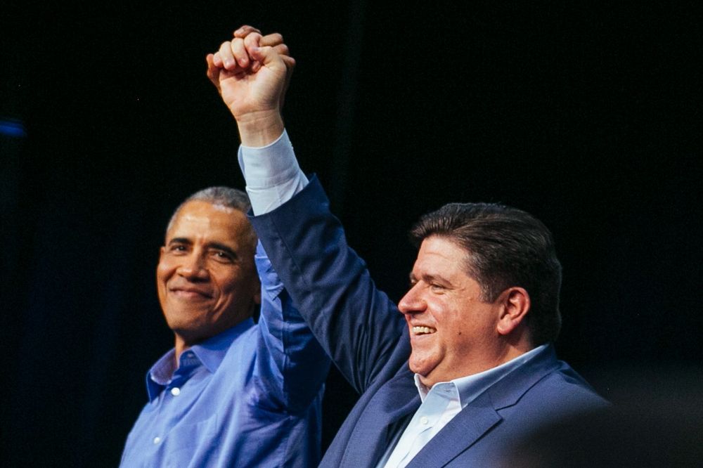 JB Pritzker and Barack Obama cheer and hold hands in the air.