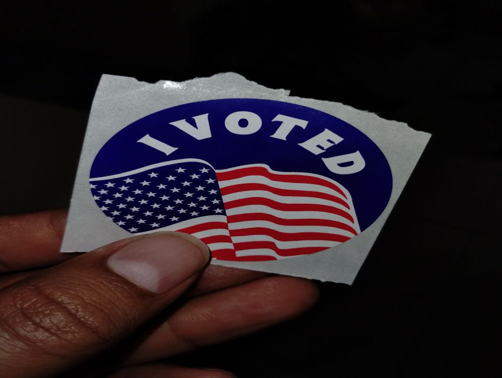 My "I Voted" sticker. I am holding the 1 inch sticker up in my hand. The words "I Voted" are in bold along the top and a waving American flag is on the bottom 