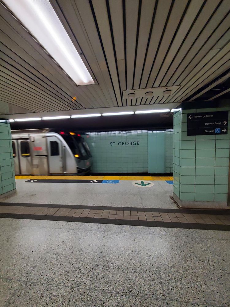 View of a train leaving the St. George subway station. The walls are tiled in dingy bluish off-mint green. The floors are institutional terrazzo. The ceiling is low, grooved dirty tan, with pallid fluorescent lighting.
