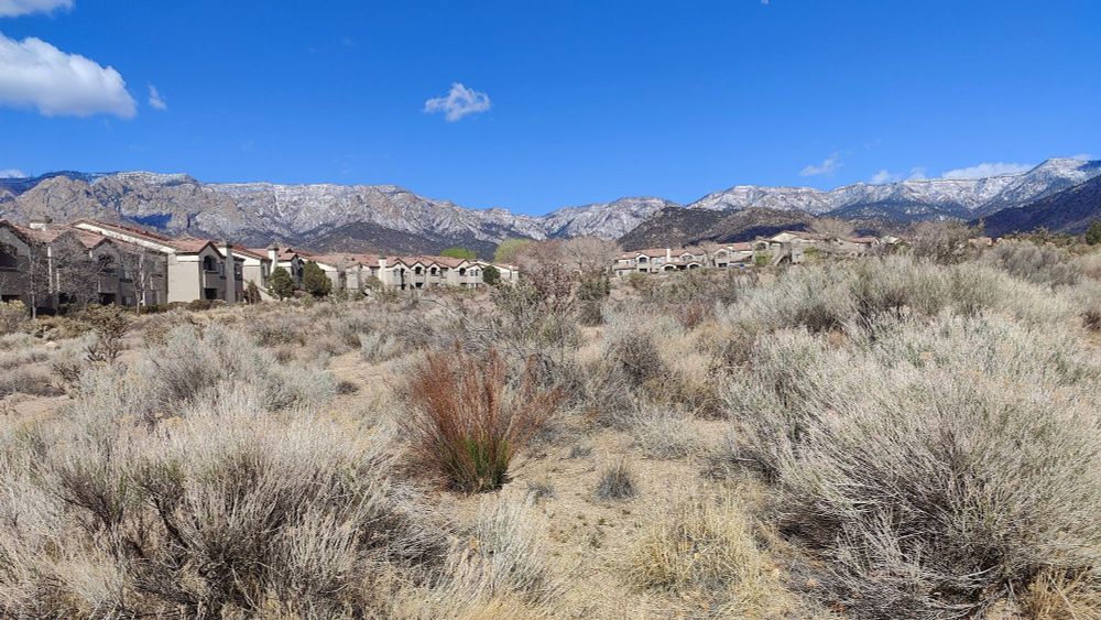Dry desert foreground, apartments mid, and snow-dusted mountains far. Blue sky, clear clouds. A perfect day.