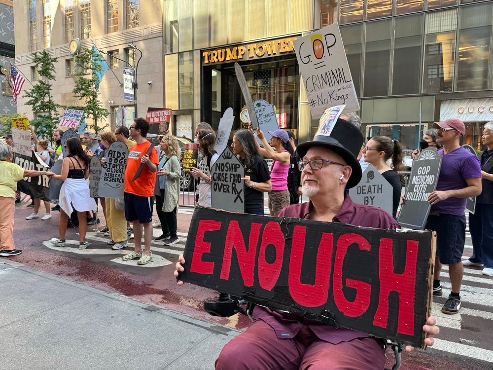 A person holding a sign that says enough in front of Trump Tower. People in background holding cardboard tombstones.