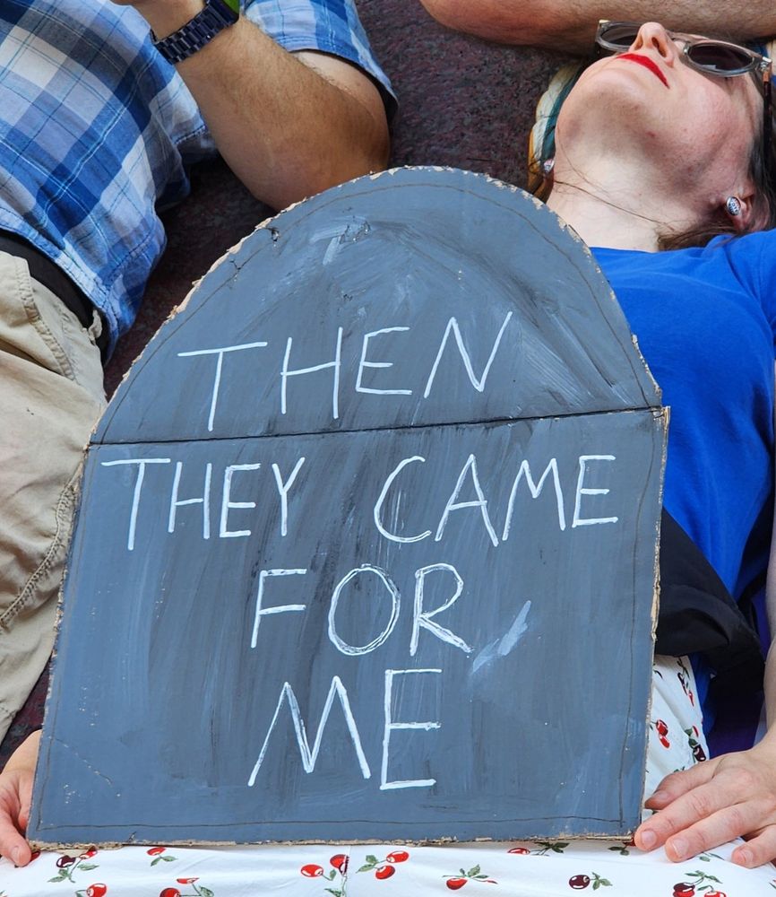 A person laying down in front of Trump tower holding a tombstone that says “then they came for me”