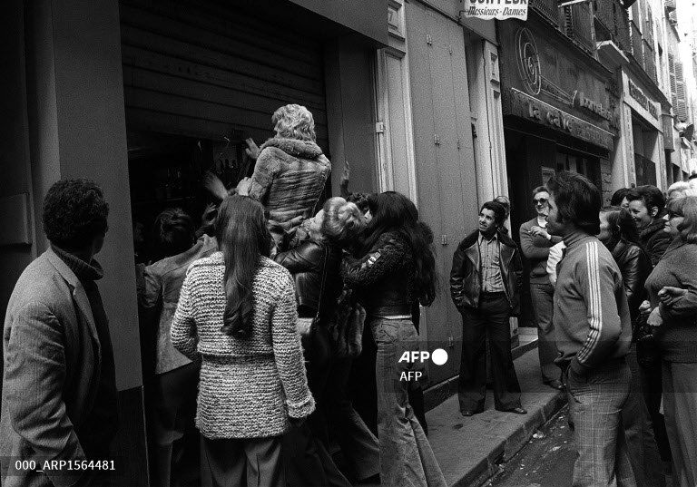 Des femmes forcent le rideau qui ferme un batiment sous le regard amusé d'hommes. Noir et blanc.