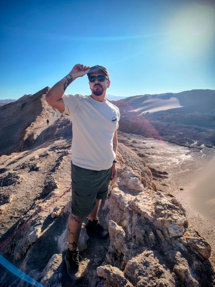 Na foto estou eu em pé (vestindo uma bermuda de trekking cor verde, camiseta creme, óculos escuto e boné), homem médio com barba. Estou sob rochas e em voltas um desfiladeiro de um deserto 