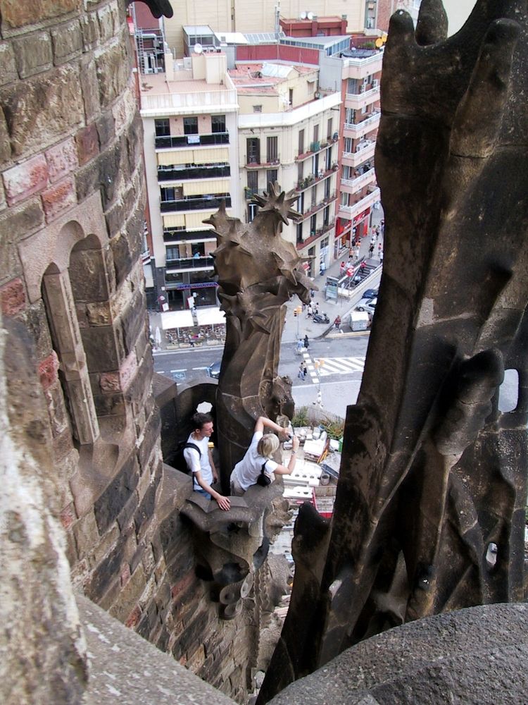 View from a balcony of one of the towers of the Sagrada Familia in Barcelona down onto another balcony, from which two other tourists, dressed in white t-shirts are taking pictures of the city from above. Some Barcelona buildings in the background.