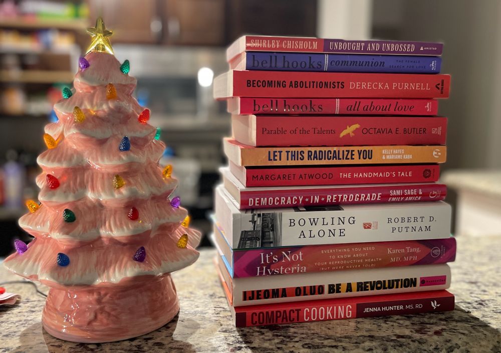 A stack of books sitting on a brown granite counter next to a small pink ceramic Christmas Tree decoration with multicolored lights. 
The books are:
Unbought and Unbossed by Shirley Chisholm
communion and all about love by bell hooks
Becoming Abolitionists by Derecka Purnell
Parable of the Talents by Octavia E. Butler
Let this Radicalize You by Kelly Hayes and Mariambe Kaba
The Handmaid’s Tale by Margaret Atwood
Democracy in Retrograde by Sami Sage and Emily Amick
Bowling Alone by Robert D. Putnam
It’s Not Hysteria by Karen Tang
Be A Revolution by Ijeoma Oluo and
Compact Cooking by Jenna Hunter