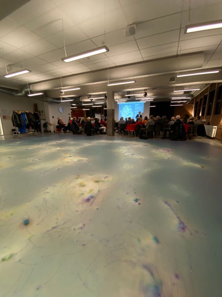 In the foreground there is a large scale data visualisation printed on a vinyl carpet. In the background there are 80 chairs with people sitting on them, looking at a stage and a screen where a presenter is giving a talk.